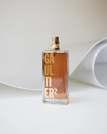 A clear glass perfume bottle with gold lettering and hardware stands on a white surface with a curved white backdrop in a studio setting.