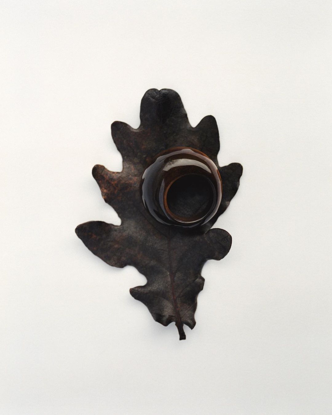 Overhead studio shot of a dark brown glazed ceramic ring holder sitting on a textured brown leaf against a clean white backdrop.