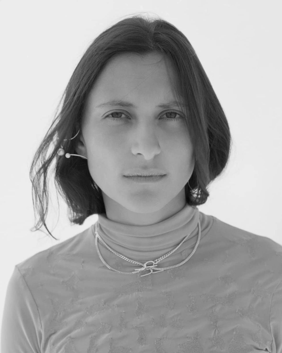 Monochrome portrait of a woman with short brunette hair wearing a turtleneck and layered necklaces against a white backdrop.