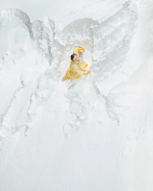 Aerial view of a woman in a yellow dress lying on a textured white surface, resembling snow or gypsum, illuminated by bright light.
