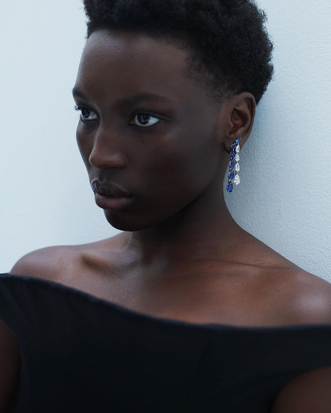 Close-up shot of a woman with dark skin and short hair, wearing blue and white dangling earrings and a black, off-the-shoulder top.