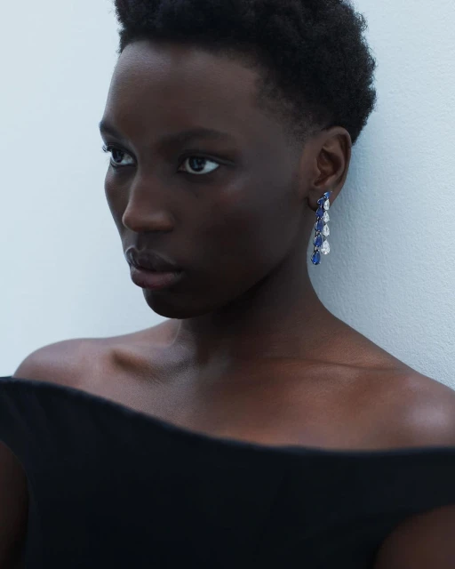 Close-up shot of a woman with dark skin and short hair, wearing blue and white dangling earrings and a black, off-the-shoulder top.