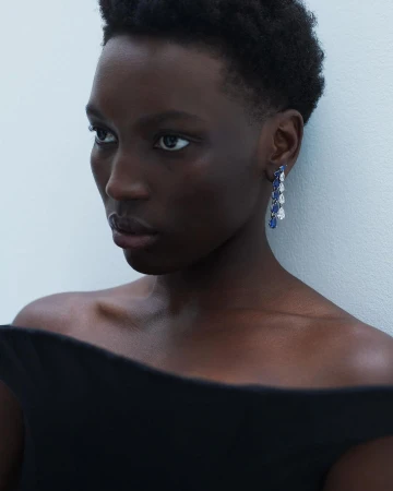 Close-up shot of a woman with dark skin and short hair, wearing blue and white dangling earrings and a black, off-the-shoulder top.