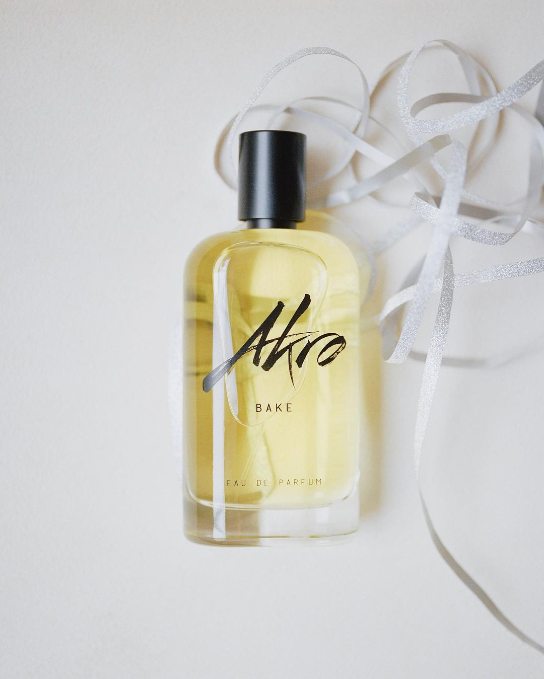 Still life studio shot of an Akro Bake perfume bottle against a white background with a decorative silver ribbon.