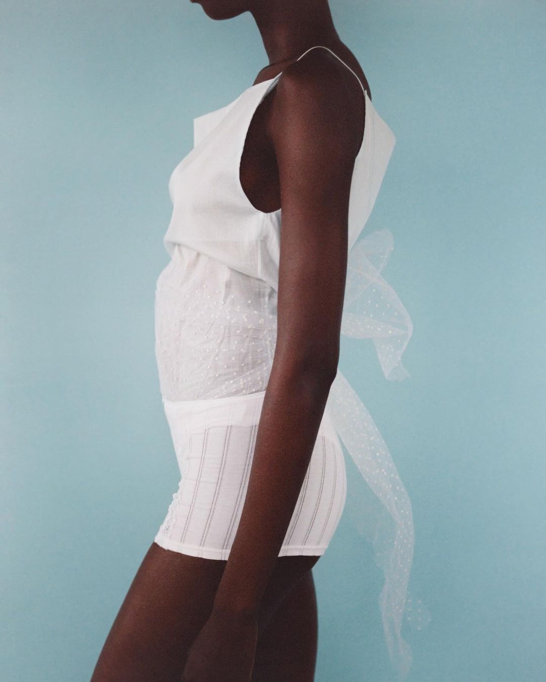 Side profile of a slim African-American woman in a white sleeveless top and shorts against a light blue backdrop, highlighting her elegant pose.