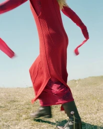 Close-up of a model in a red knit dress and dark boots walking across an outdoor grassy terrain under a blue sky.