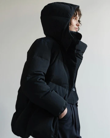 Model wearing a black hooded puffer jacket and matching trousers against a plain light wall in Paris by Aude Le Barbey.