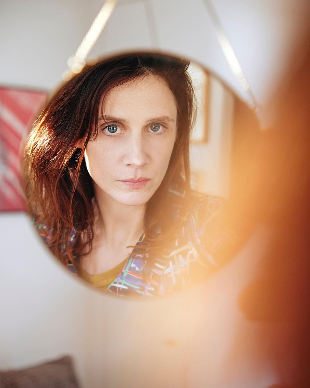 Close-up portrait of a woman with blue eyes, reflected in a round mirror indoors, with natural lighting and an orange blurry foreground element.
