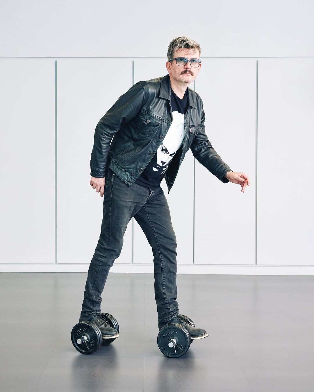 Adult male with a mustache in a black jacket and jeans is standing on custom roller skates with weights in a bright, minimalistic indoor space.