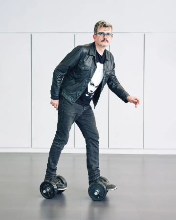 Adult male with a mustache in a black jacket and jeans is standing on custom roller skates with weights in a bright, minimalistic indoor space.