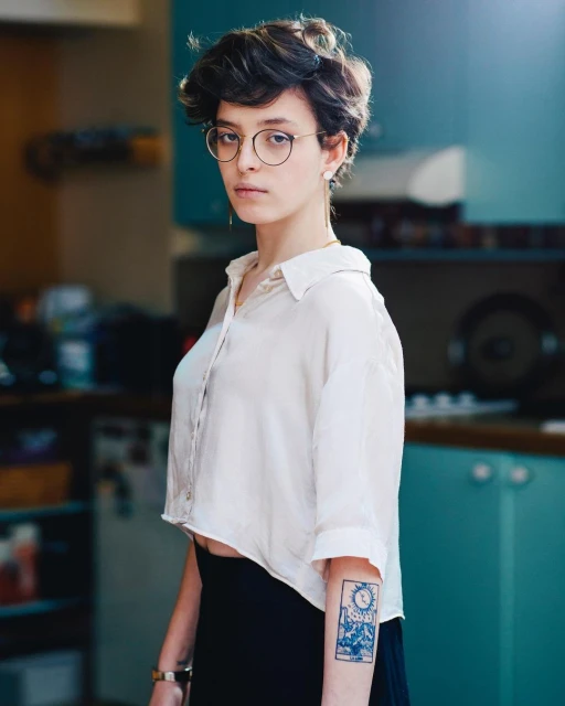 Indoor portrait of a young adult female with short brown hair wearing glasses, a white blouse and a black skirt in front of a kitchen area.