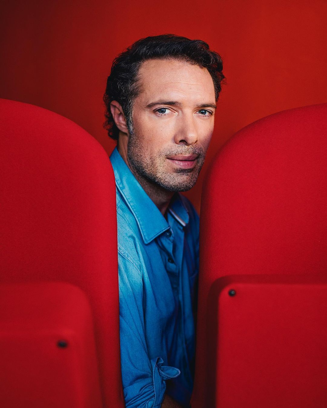 Portrait of an adult male wearing a blue shirt, seated between two red chairs, set against a red backdrop in a photo studio.