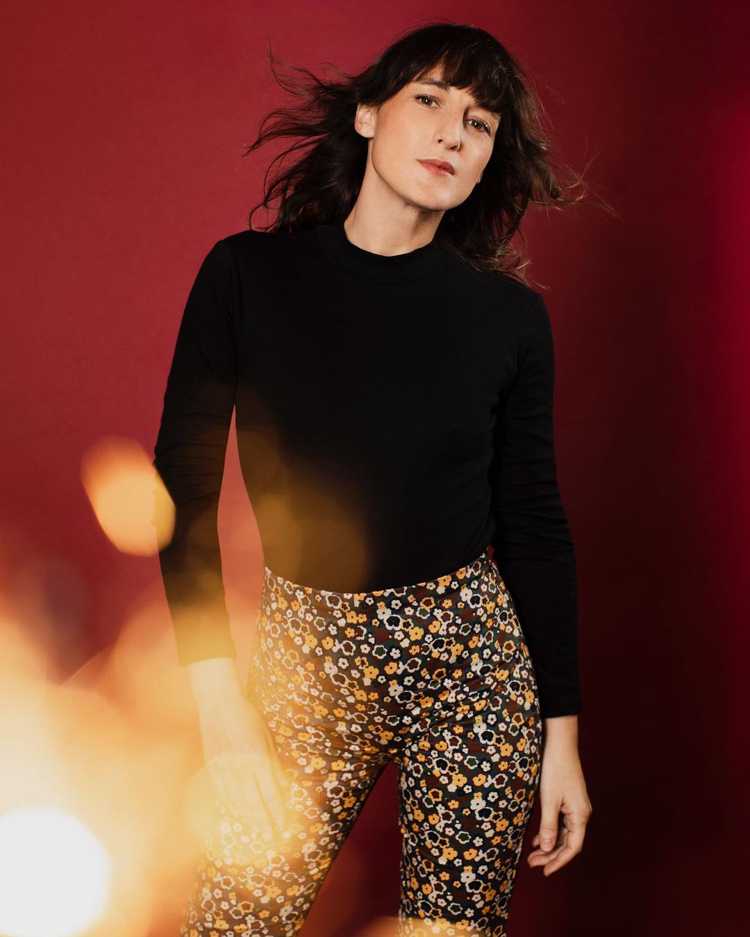 Portrait of a young adult woman with dark hair, wearing a black top and patterned pants, posing against a red backdrop in a studio.