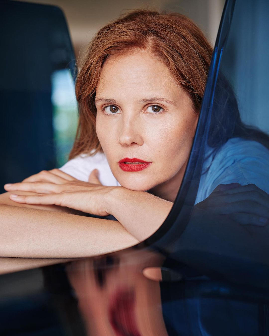 Portrait of a red-haired woman with red lipstick, leaning on her arms indoors, with a reflective surface and a blue panel.