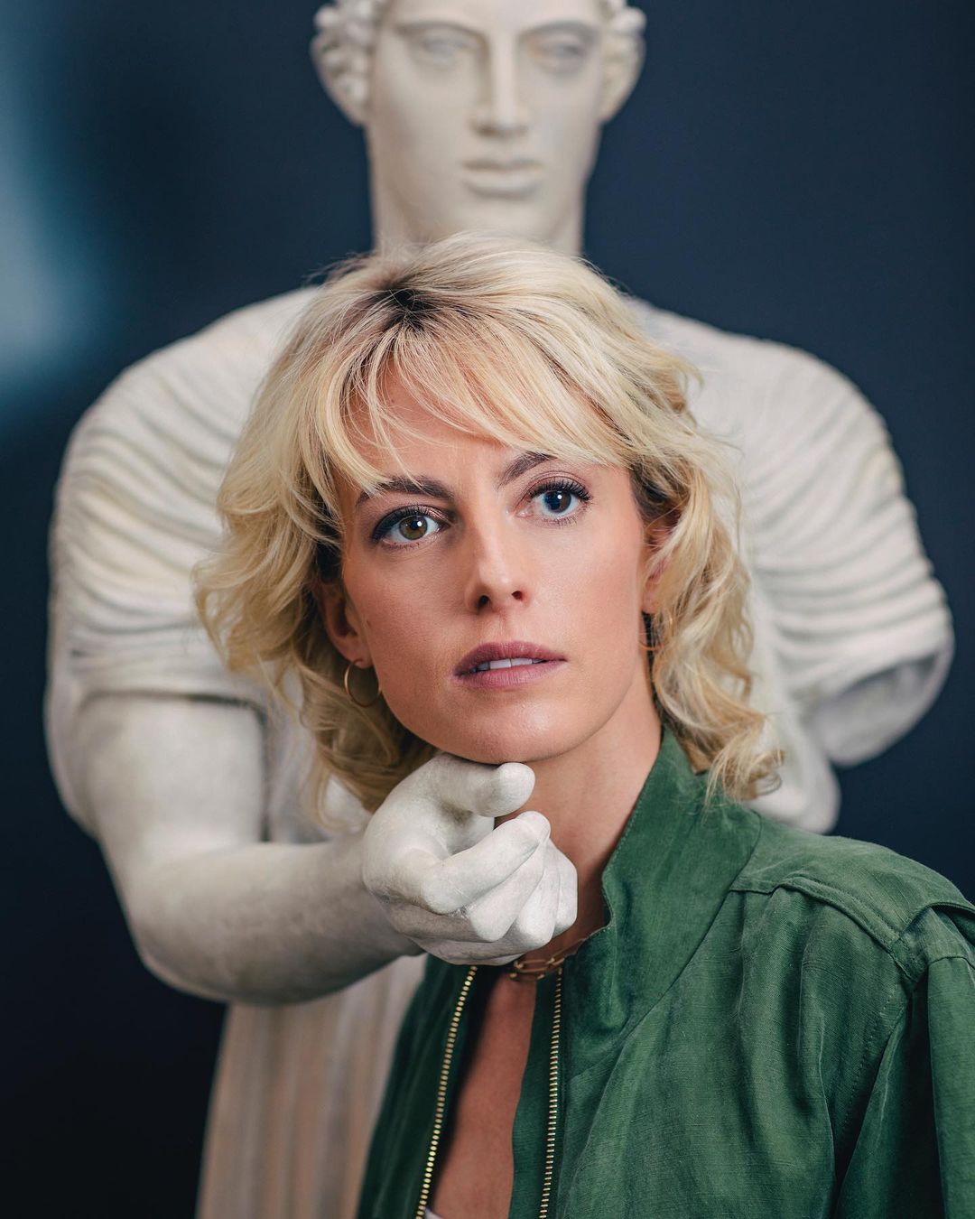 A woman with heterochromia stands with a white marble statue, the statue's hand near her neck, in a portrait-style shot, indoor.