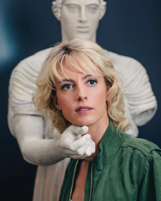 A woman with heterochromia stands with a white marble statue, the statue's hand near her neck, in a portrait-style shot, indoor.