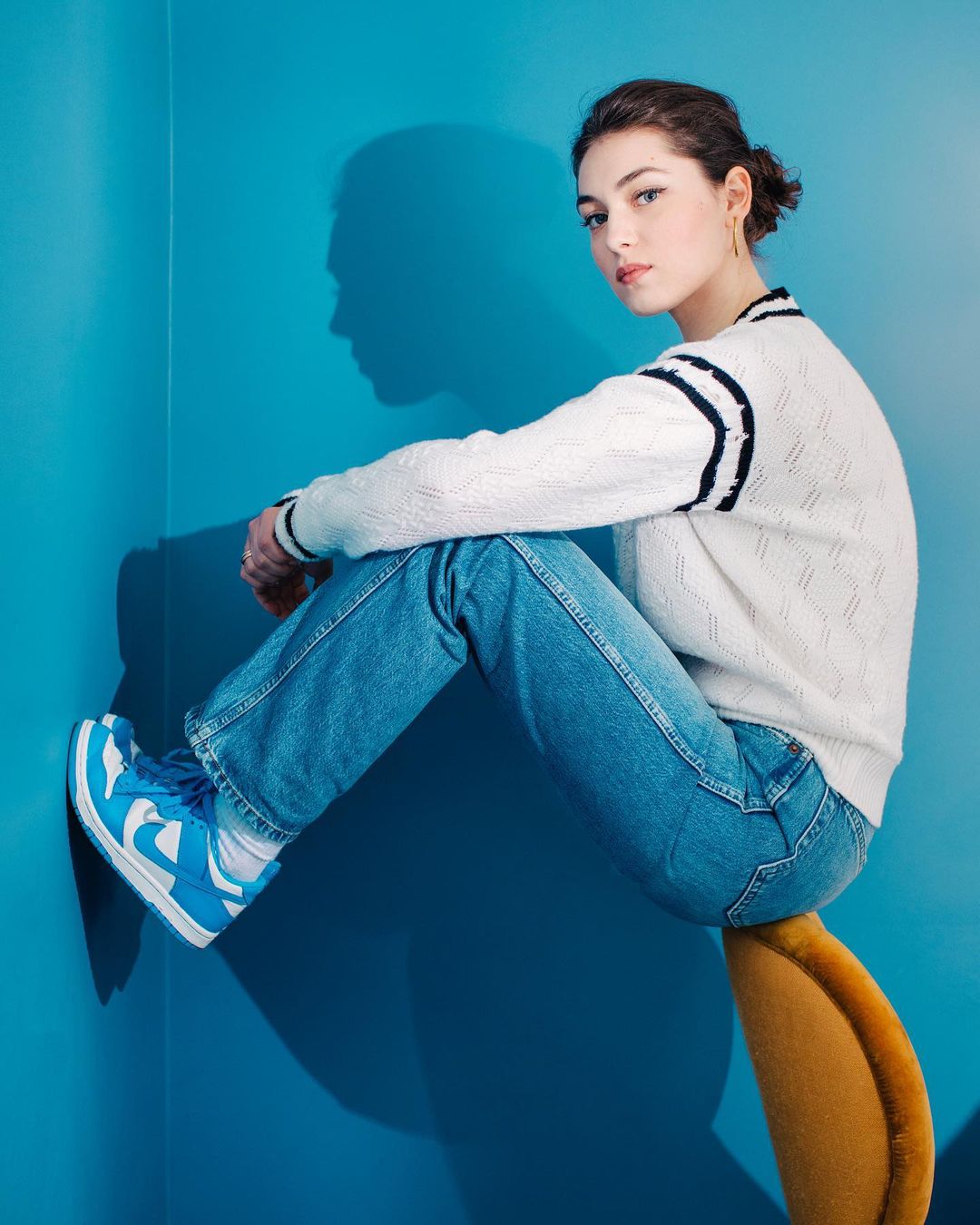 Young woman in blue jeans and blue and white sneakers sits on a chair against a blue wall in a casual fashion pose.