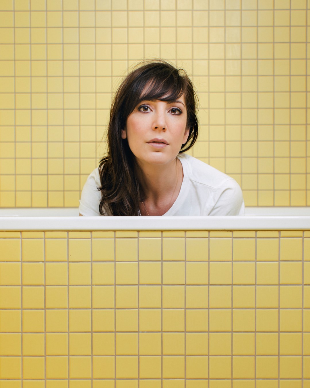 Woman in a bathtub with yellow tiled walls. Portrait shot of a brunette in a white shirt looking at the camera in an indoor setting.