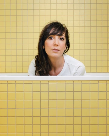 Woman in a bathtub with yellow tiled walls. Portrait shot of a brunette in a white shirt looking at the camera in an indoor setting.