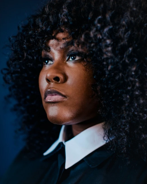 Close-up studio portrait of an African-American woman with curly hair wearing a black collared shirt looking upward and left, with a dark blue background.
