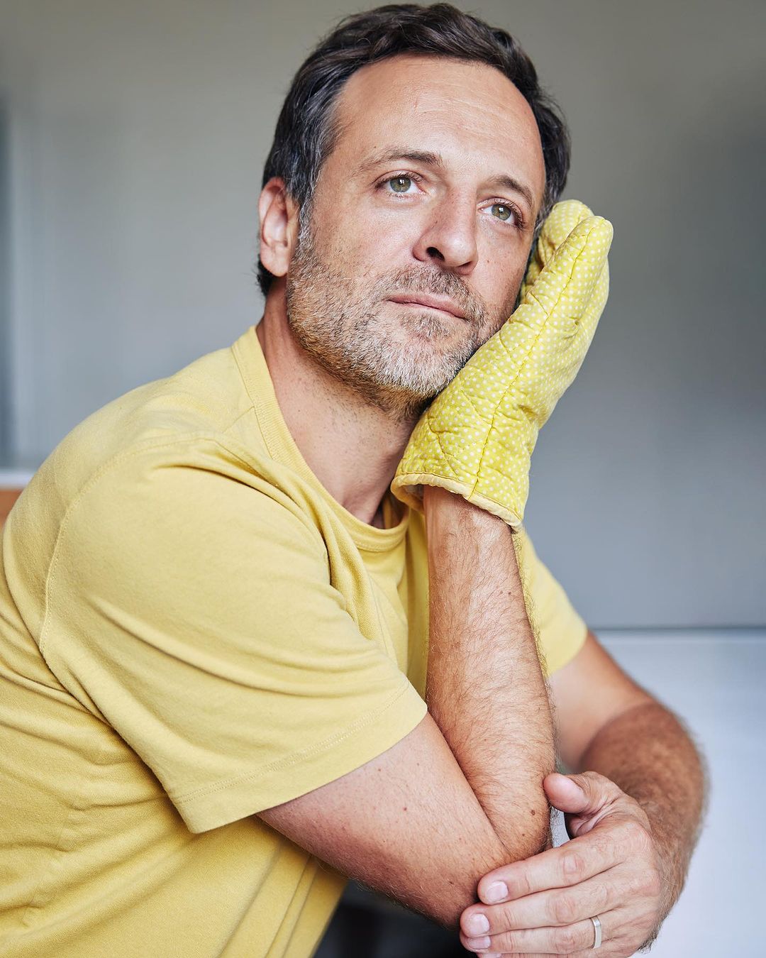 Portrait of a man resting his head on his hand, which is covered with a yellow oven mitt, with a grey background.