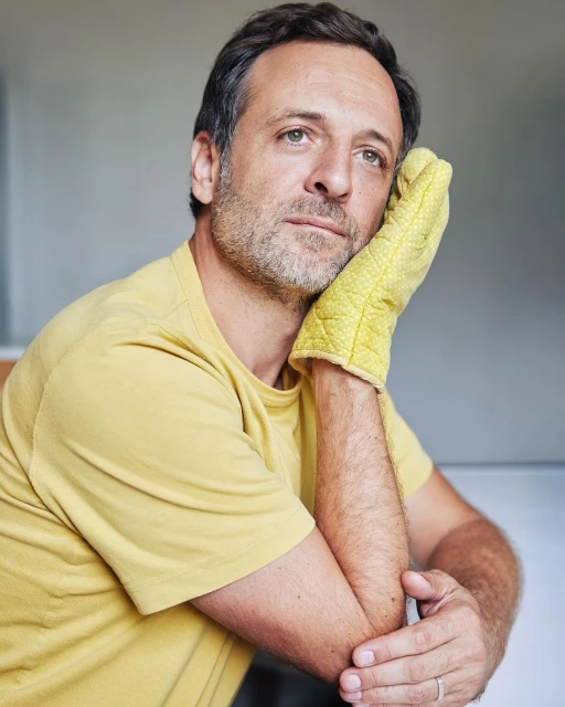 Portrait of a man resting his head on his hand, which is covered with a yellow oven mitt, with a grey background.