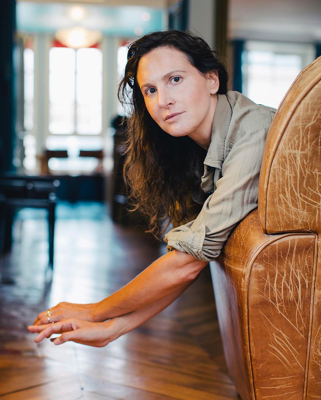 Portrait of a woman with long brown hair reclining in a leather chair, looking at the camera, indoors with wood floors.