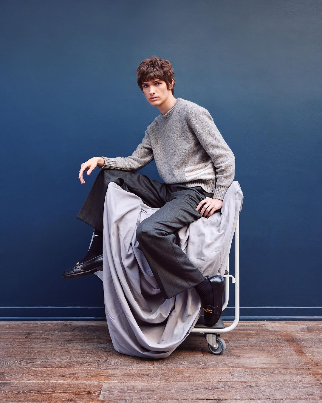 A stylish young man in a gray sweater and dark trousers poses while seated in a laundry cart against a deep blue backdrop.
