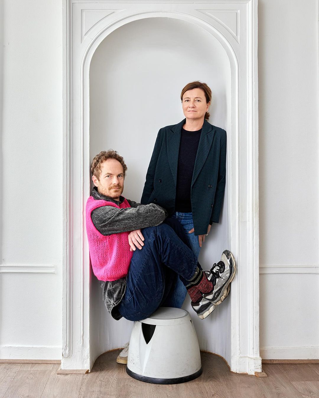 Indoor portrait of a man sitting on a stool and a woman standing inside of a white archway in a minimalist, light-filled interior.