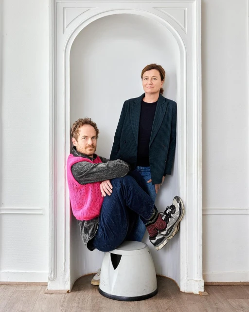 Indoor portrait of a man sitting on a stool and a woman standing inside of a white archway in a minimalist, light-filled interior.