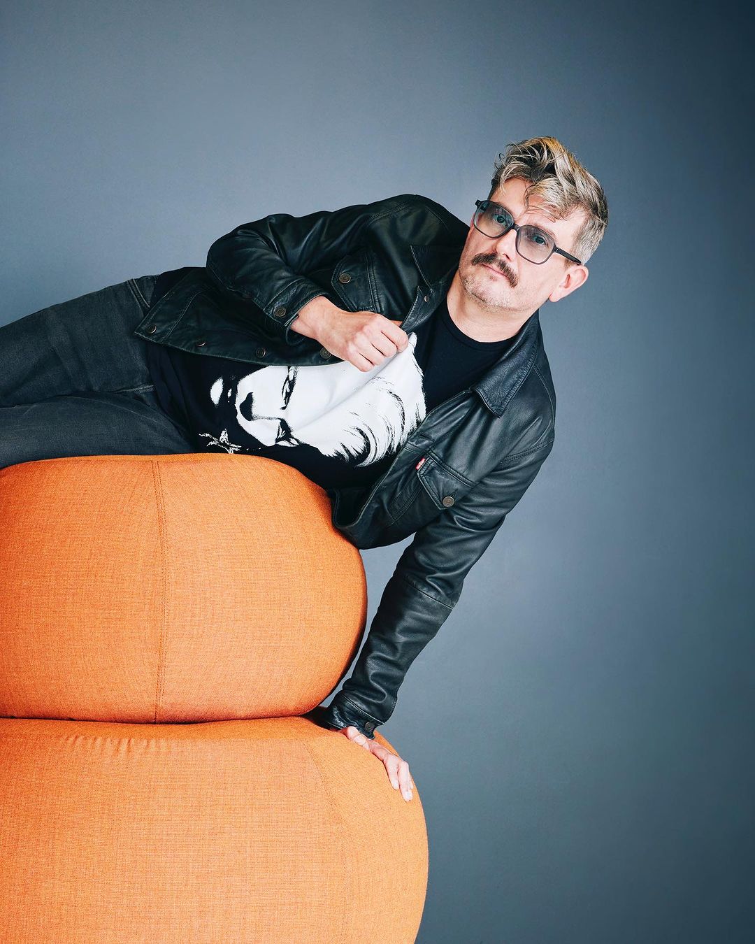 Adult male in black jacket and glasses reclining on an orange pouf in a studio setting, looking at the camera.