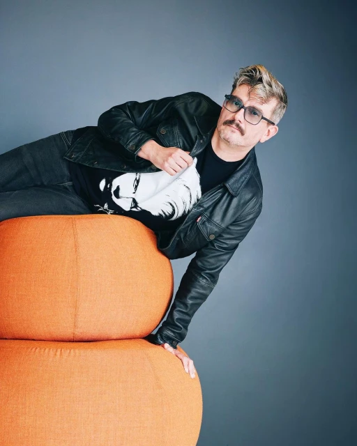 Adult male in black jacket and glasses reclining on an orange pouf in a studio setting, looking at the camera.