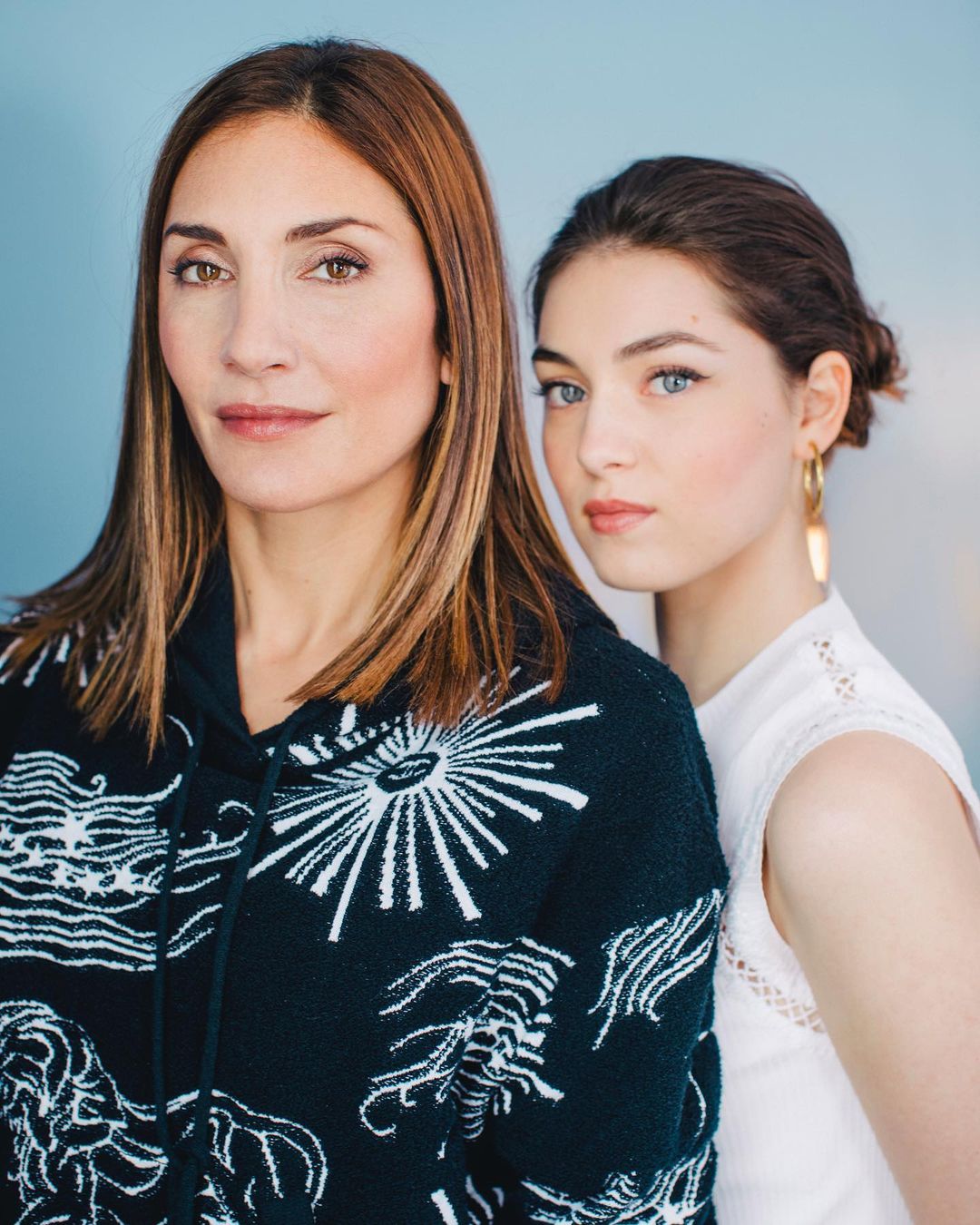 Portrait of an adult woman with long brown hair and a teenage girl with gold earrings, both posing indoors with a light blue backdrop.