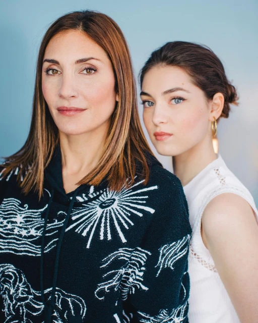 Portrait of an adult woman with long brown hair and a teenage girl with gold earrings, both posing indoors with a light blue backdrop.