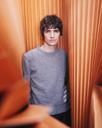 Indoor portrait of a young adult male with dark hair wearing a grey sweater in front of an orange pleated background. Casual style.