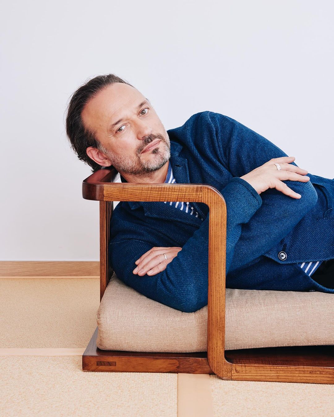 Adult man reclining on a modern wood and tan woven chair, wearing a blue blazer and striped shirt, against a white background.