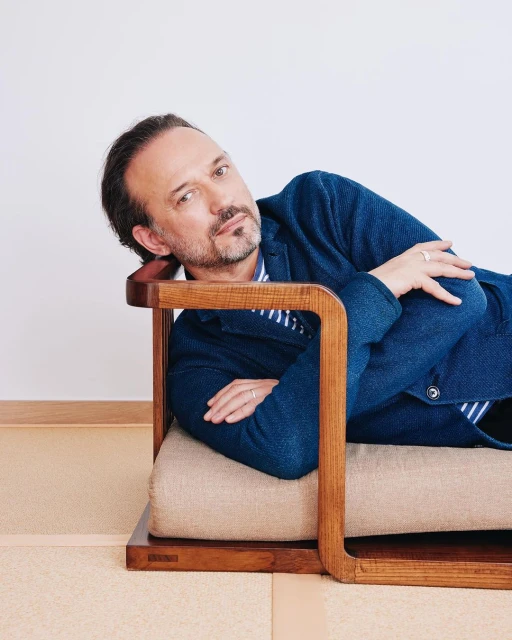 Adult man reclining on a modern wood and tan woven chair, wearing a blue blazer and striped shirt, against a white background.