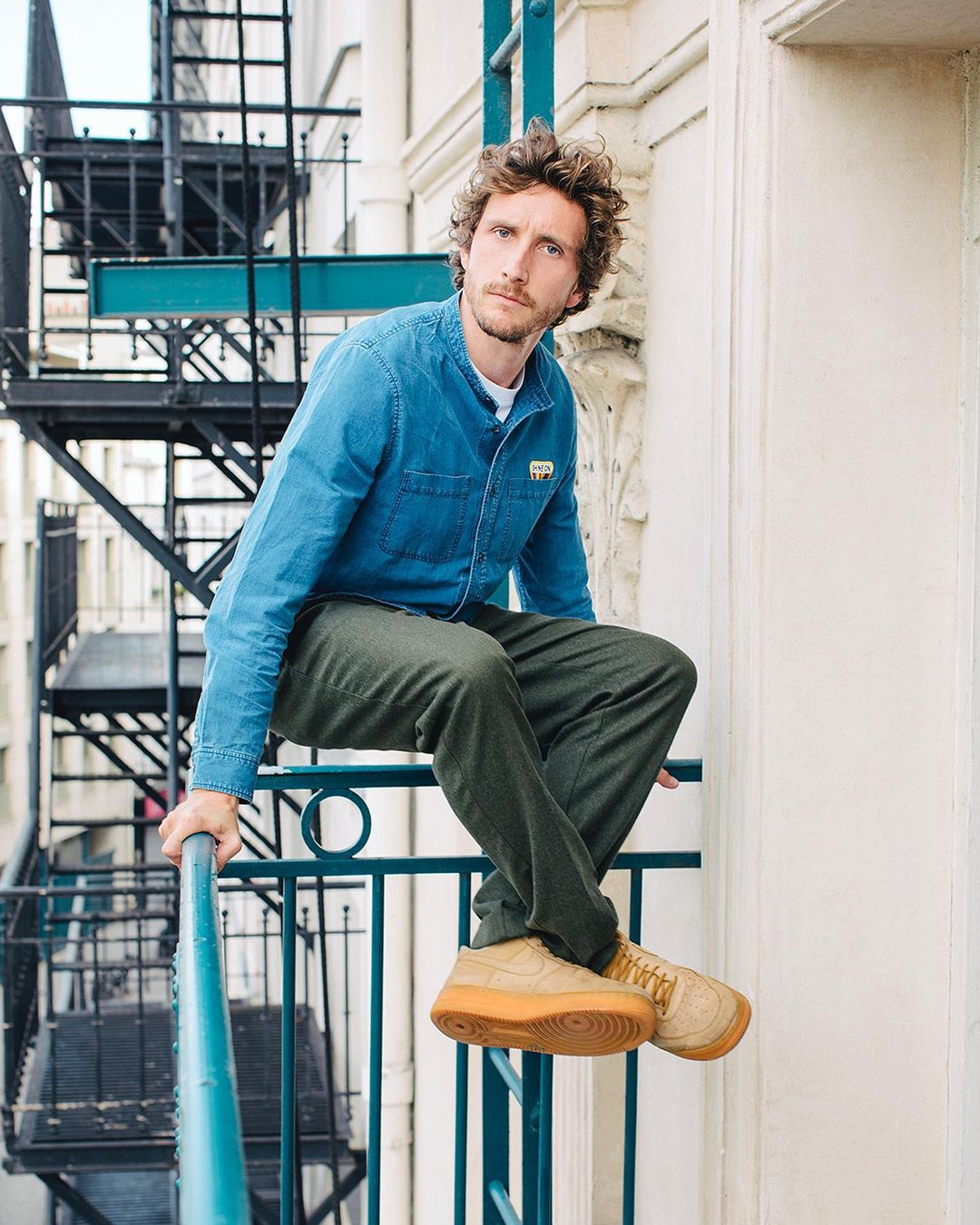 Adult male poses on a turquoise railing outside. He is wearing a denim shirt, khaki pants, and tan sneakers in an urban environment.