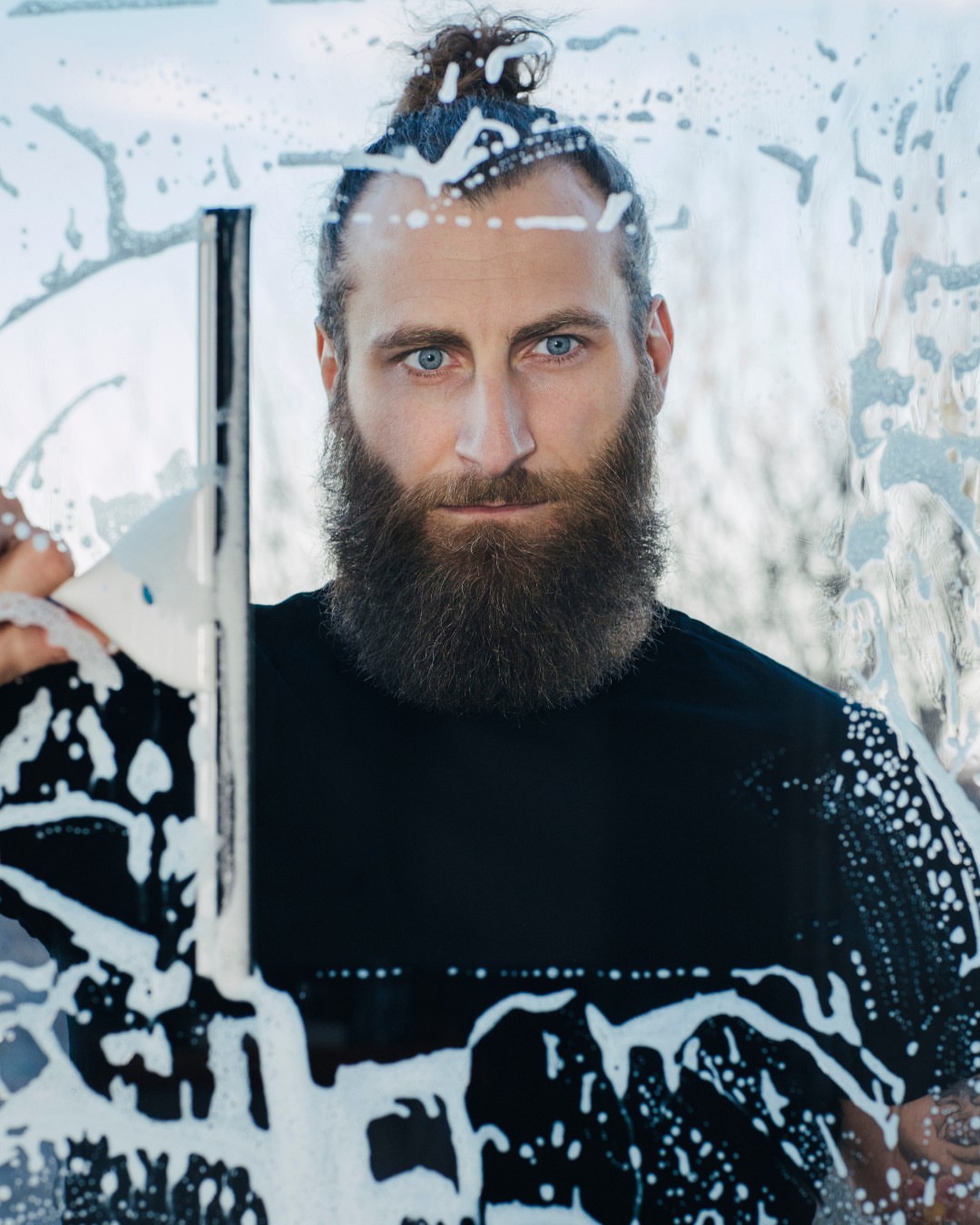 Adult man with beard and top knot cleaning window with squeegee, creating abstract patterns of white soap suds on glass.