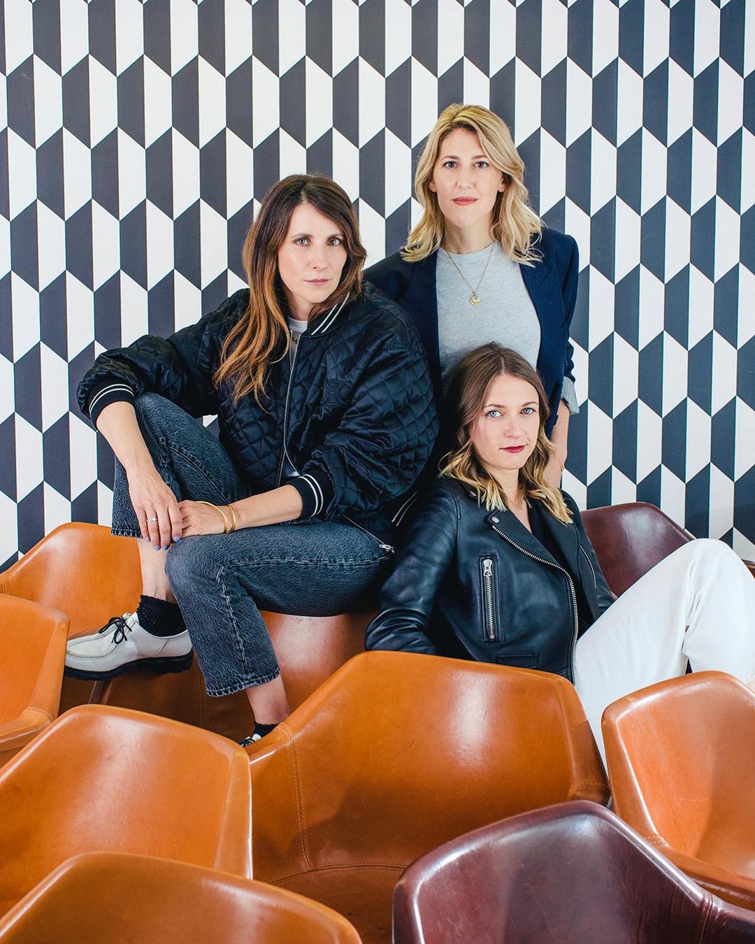 Three adult women pose on and around a collection of tan leather chairs in front of a geometric black and white patterned wall.