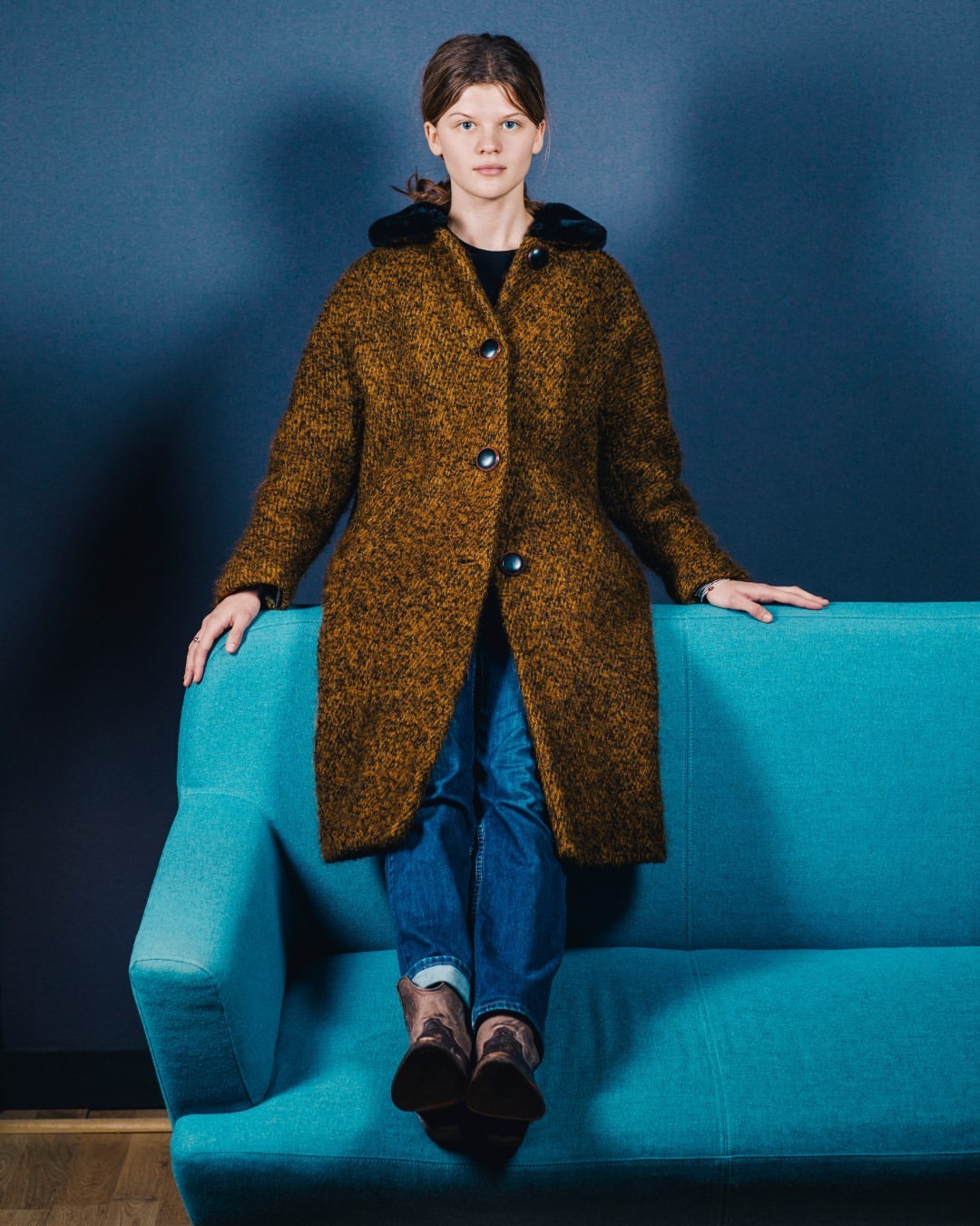A woman in a brown coat sits on a turquoise couch in a studio. The image captures a fashion-forward look in a posed portrait.