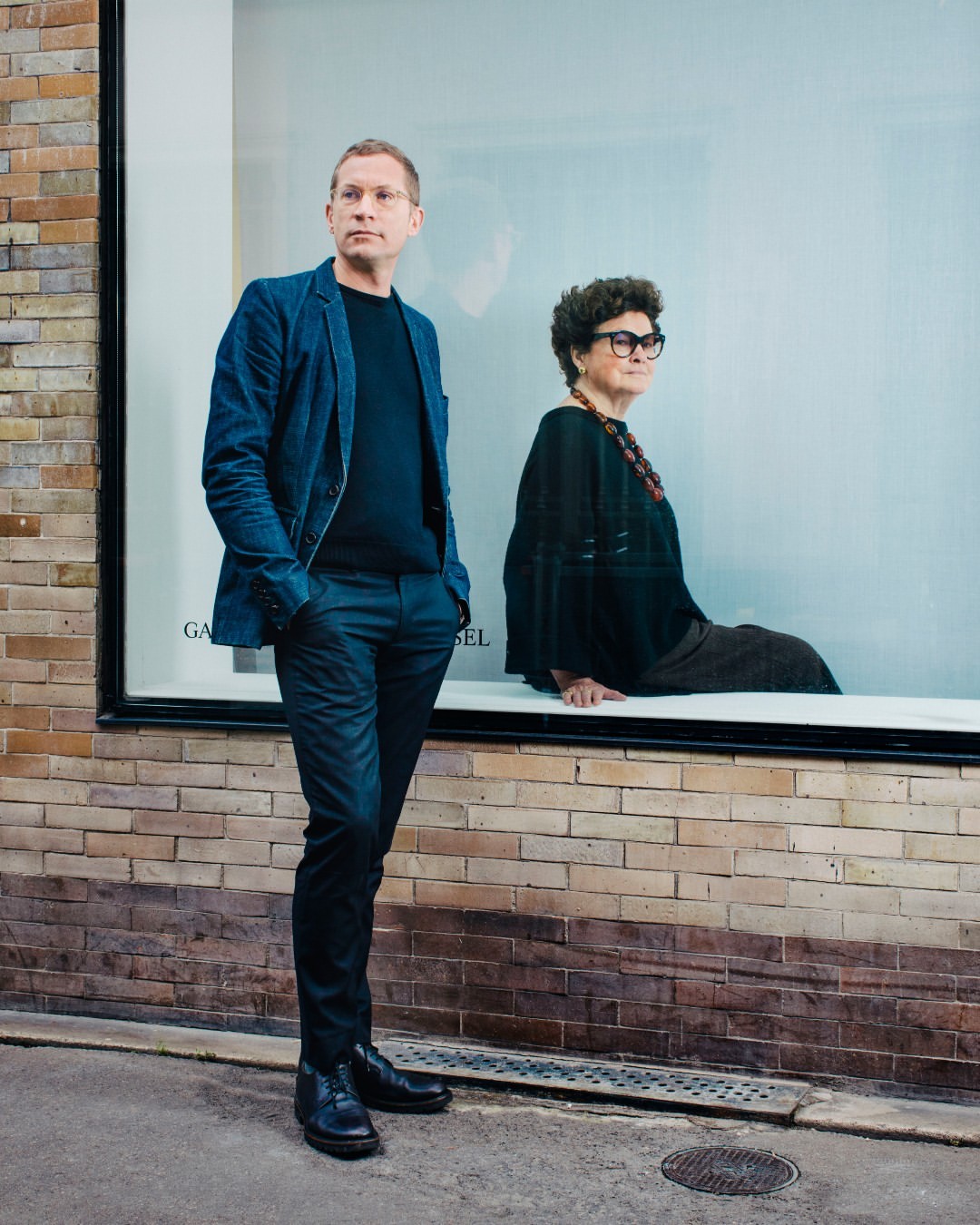Man in a blue jacket and woman with glasses pose in front of a brick building with a window in an urban setting on an overcast day.
