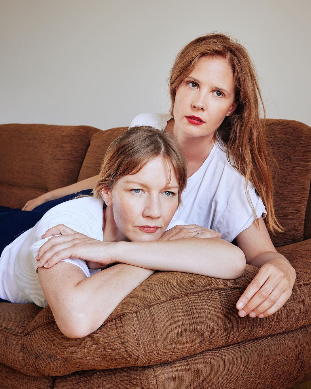 Two adult women pose closely together indoors on a brown couch. One woman is lying down, with the other woman behind her.