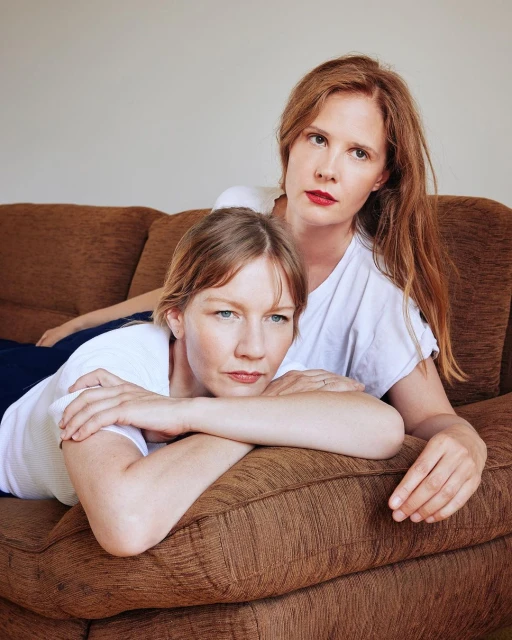 Two adult women pose closely together indoors on a brown couch. One woman is lying down, with the other woman behind her.