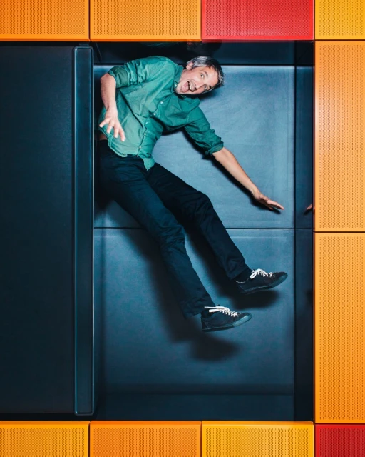 Adult man laying in a box with orange and red wall panels, wearing green shirt and black pants, with a smiling facial expression.