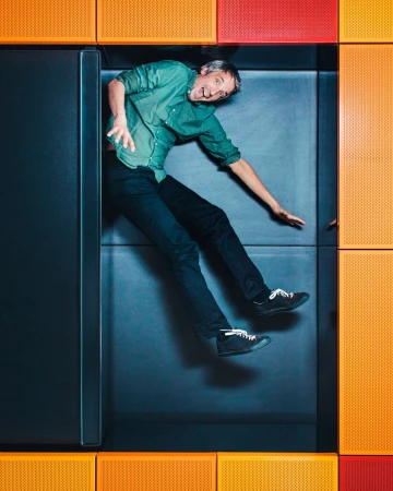 Adult man laying in a box with orange and red wall panels, wearing green shirt and black pants, with a smiling facial expression.