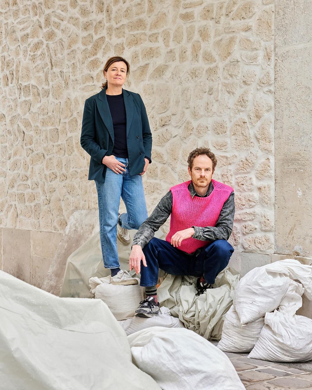 Two adult people outdoors. One woman stands near a man squatting on bags, set against a stone wall, captured in natural light.