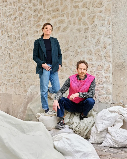 Two adult people outdoors. One woman stands near a man squatting on bags, set against a stone wall, captured in natural light.