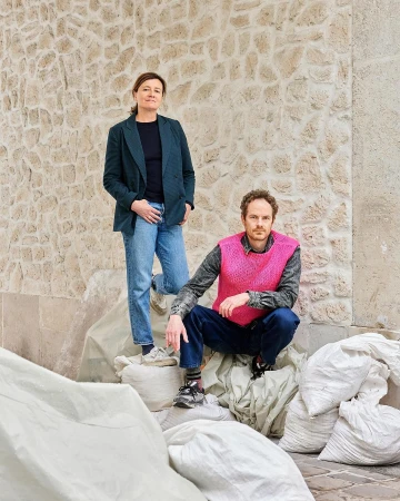 Two adult people outdoors. One woman stands near a man squatting on bags, set against a stone wall, captured in natural light.