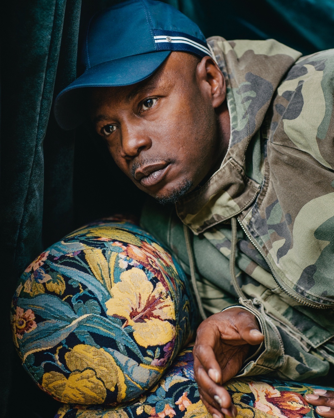 Portrait of an adult male wearing a camouflage jacket and baseball cap, sitting on an armchair with a floral upholstery.